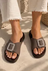 Sandalias planas color marrón con suela de aspecto amaderado y plantilla negra con logo dorado. Poseen una tira ancha de gamuza en el empeine decorada con una hebilla rectangular grande con pedrería brillante.