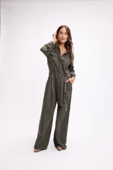 Mono de estilo utilitario color beige, con cierre frontal de botones, bolsillos en el pecho y cintura entallada con detalle de cadenas decorativas. Presenta mangas largas y piernas de corte recto y holgado.