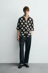 Camisa de manga corta con cuello solapa y cierre frontal de botones, confeccionada en tejido de crochet con patrón de flores en negro y beige.