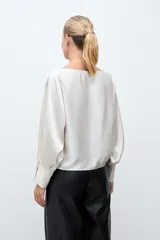 Blusa de satén color crudo, con cuello bote, mangas largas con aberturas laterales y puños abotonados.