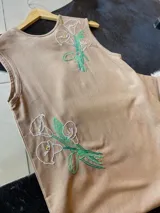 Vestido midi color beige confeccionado en lino de algodón, con diseño sin mangas y cuello redondo. Presenta bordados artesanales a mano de ramos de flores en color blanco y verde en el frente.