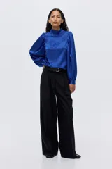 Blusa de tejido jacquard con acabado satinado y diseño floral en relieve. Presenta cuello elevado, mangas largas abullonadas con puños abotonados y cierre posterior con botones forrados.