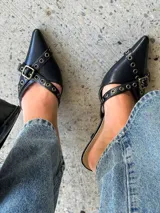 Mules de cuero negro con punta fina, diseño abierto en los laterales y tiras decorativas con ojales metálicos y hebilla ajustable.