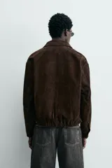 Cazadora de hombre de corte relajado confeccionada en piel con acabado ante, color marrón oscuro. Presenta cuello solapa, manga larga con puños abotonados a presión, bolsillos de vivo en cadera, bolsillo interior y bajo con elástico. Cierre frontal oculto por solapa con botones a presión.
