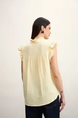 Camisa color amarillo pálido con textura arrugada, cuello mao con volados, cierre frontal con botones y mangas cortas con volados.