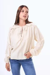 Blusa de satén color beige, con cuello redondo, detalle de volados en los hombros y cierre frontal con lazo. Presenta mangas largas con puños elásticos.