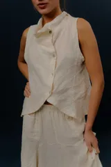 Chaleco de lino color marfil o beige claro, con diseño sin mangas, cuello alto amplio, cierre frontal con botones y bolsillos delanteros amplios. Presenta un corte asimétrico y relajado.