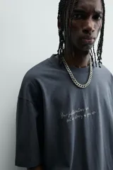 Camiseta de algodón color gris oscuro, corte relajado (relaxed fit), con cuello redondo y manga corta. Presenta dos inscripciones bordadas en hilo blanco en el frente.