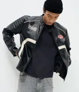 Campera biker de cuero sintético negro con detalles en blanco, cuello mao con broche, cierre frontal y parches bordados en el pecho. Presenta diseño acolchado en los hombros y mangas largas.