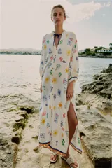 Vestido largo estilo túnica de color crudo con estampado multicolor de motivos marinos y mediterráneos. Presenta cuello con volante, cierre frontal con botones, manga larga y cordón ajustable en el cuello con abalorios.