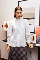 Camisa de manga larga con estampado de rayas verticales celestes sobre fondo blanco. Presenta un cuello alto con volados, cierre frontal con botones y puños con volados.