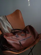 Bolso de viaje tipo weekender de cuero color tostado con asas y detalles en marrón oscuro, herrajes metálicos y bolsillo frontal con cierre.