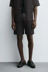 Bermuda de hombre, corte regular fit, color oscuro con patrón de rayas verticales finas en tono marrón claro. Presenta cintura elástica ajustable con cordón interior, bolsillos frontales y un bolsillo trasero de plastrón.