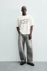 Camiseta blanca de algodón con corte boxy fit y cuello redondo. Presenta un estampado frontal con texto manuscrito en negro y marrón.