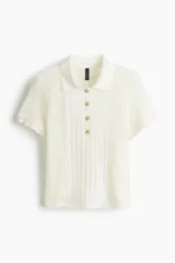 Remera polo entallada de color blanco, confeccionada en punto calado suave. Presenta cuello clásico, tapeta corta con botones frontales, panel central en punto acanalado y mangas cortas con ribete festoneado.