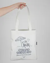 Bolso de mano tipo tote bag color crudo, con asas largas y estampado con motivos veraniegos en azul: sombrilla, reposera, perro caniche y texto "Verano Uruguayo".
