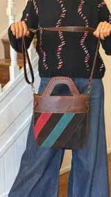 Bolso tipo matera confeccionado en cuero vacuno, con diseño estructurado y manija superior integrada. Presenta un cuerpo con franjas diagonales en gamuza de colores contrastantes (amarillo, verde y azul) sobre una base de cuero marrón, con detalles de costuras artesanales en los bordes.