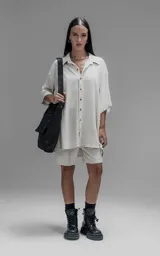Camisa de corte oversize con cuello camisero, cierre frontal de botones y mangas cortas. Presenta un diseño holgado con bolsillos laterales integrados en la costura.