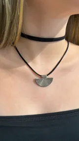 Gargantilla doble compuesta por un choker de cinta negra lisa y un collar de cordón negro con dije metálico plateado en forma de semicírculo con diseño de líneas concéntricas.