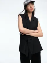 Chaleco sastrero negro de silueta estructurada, con solapas clásicas y lazo ajustable en la cintura. Presenta un diseño sin mangas con bolsillos laterales integrados.