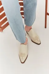Mule de cuero color suela con diseño de punta fina y bordado decorativo en el empeine. Presenta un taco bajo de madera y una silueta abierta en el talón.