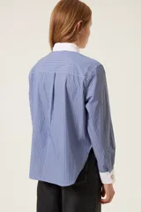 Camisa de popelina a rayas azules y blancas, de corte recto, con cuello y puños blancos. Presenta un babero plisado blanco.