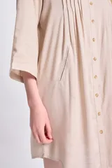 Vestido camisero corto de color beige, con cuello clásico, cierre frontal de botones y detalle de tablas verticales en el frente. Presenta mangas tres cuartos y un corte holgado con ruedo redondeado.