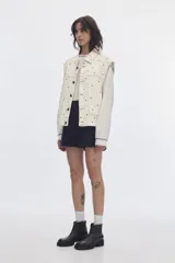 Chaleco oversize de denim color crudo con estampado de lunares negros, cuello camisero, bolsillos delanteros y cierre frontal de botones metálicos.