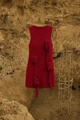 Vestido corto de tejido de punto color rojo con destellos de lúrex, sin mangas y cuello a la base. Presenta apliques decorativos en forma de flores de tela con tiras colgantes distribuidos a lo largo de la prenda.