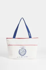 Bolso tipo shopper color blanco con detalles en rojo, confeccionado en algodón. Presenta un estampado central en azul con el escudo nacional y la frase 'El País de los mates y las siestas'. Cuenta con correas de hombro en color azul.