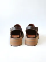 Zueco de cuero gamuzado color chocolate, con diseño de punta cerrada y tira ajustable con hebilla metálica en el talón. Presenta una suela de plataforma dentada en tono marrón claro.