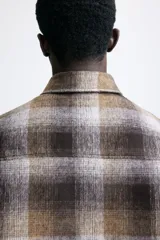 Sobrecamisa acolchada de corte relajado con diseño a cuadros en tonos marrones, grises y negros. Presenta cuello inglés, cierre frontal con botones a presión, hombros caídos y mangas largas con puños abotonados.