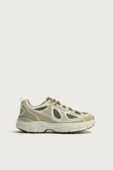 Zapatilla deportiva tipo running con diseño de suela voluminosa e irregular. Presenta una combinación de piezas en tonos beige y crema, con paneles de malla y cordones jaspeados.