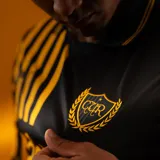 Camiseta de fútbol Puma del Club Atlético Peñarol, edición aniversario, modelo de manga larga. Diseño con mitad derecha a rayas verticales amarillas y negras, y mitad izquierda negra con escudo del CURCC y logo de Puma en dorado. Presenta cuello polo negro y sponsor antel en el pecho.