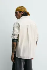 Camisa blanca de algodón con corte relajado, cuello solapa, manga larga con puño abotonado y bolsillo de plastrón en el pecho. Presenta bajo asimétrico con aberturas laterales y cierre frontal de botonadura.