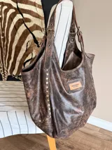 Cartera tipo tote bag marca Antonia, elaborada en cuero marrón con efecto desgastado, diseño amplio y tiras de hombro ajustables. Cuenta con detalles de tachas metálicas en los laterales y parche frontal con el logo de la marca.
