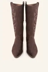 Bota de caña alta estilo tejano confeccionada en gamuza color marrón oscuro. Presenta detalles de costuras decorativas en forma de llama en la caña, taco cuadrado de madera y punta almendrada.