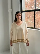 Sweater de punto color crudo con cuello en V, mangas largas y diseño holgado. Presenta detalles de franjas horizontales en color beige en los puños y en el ruedo inferior.