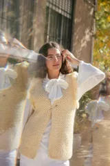 Chaleco de crochet tejido en punto cesta color crema, con diseño sin mangas y detalle de flecos en los hombros. Presenta un cierre frontal con alfiler de gancho metálico.