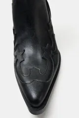 Bota estilo cowboy de caña alta, color negro, con detalles en relieve y costuras decorativas. Presenta punta fina, tiradores laterales y taco cuadrado inclinado.
