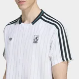Remera blanca con finas rayas verticales celestes, cuello redondo negro con ribete verde y mangas cortas con ribete negro. Presenta las tres tiras negras características de Adidas en los hombros, el logo de Adidas bordado en el pecho y el escudo del Liverpool FC bordado en el pecho.