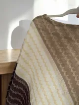 Poncho de hilo con diseño de franjas diagonales en tonos degradados de marrón, crema y beige, con textura tejida y terminación de flecos en el borde.