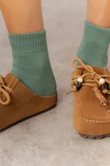 Zueco de cuero gamuzado color tostado, con cordones decorativos que incluyen cuentas de madera y detalles envueltos en cuero. Diseño abierto en la parte trasera y suela liviana.