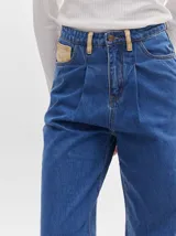 Pantalón jean de tiro alto y corte pinzado, con perneras de caída suelta y levemente anchas en el bajo. Posee pasador con hebilla en la parte trasera.