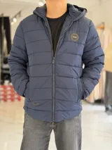 Campera puffer azul marino con capucha ajustable, cierre frontal y bolsillos laterales con cierre a presión. Cuenta con parche redondo con la inscripción "TURK" en el pecho.