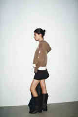 Sudadera marrón con capucha ajustable con cordones, manga larga con puños y bajo elástico. Presenta un estampado ovalado en el pecho con el texto "Texas USA" en rosa y azul, y un diseño de estrellas blancas en las mangas. El bolsillo canguro frontal tiene detalles de tachas y bordes deshilachados.