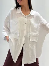 Camisa blanca de lino con corte oversize, cuello clásico, cierre frontal con botones, mangas largas con puños abotonados y dos bolsillos de parche en el pecho.