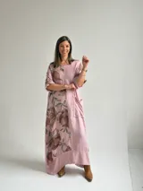 Vestido largo color rosa, con mangas cortas y diseño holgado, presenta un estampado floral en tonos marrones y verdes en un lateral, además de un bolsillo aplicado en la parte frontal y costuras visibles de estilo rústico.