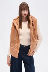 Chaleco con capucha, diseño bicolor en tonos tostado y camel, con paneles de textura de corderito y tejido técnico liso. Presenta cierre frontal con botones a presión y cordones ajustables en la capucha.