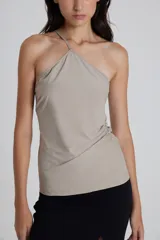 Top asimétrico color beige, con escote halter y bretel cruzado en un hombro. Presenta frunces laterales y silueta ajustada al cuerpo.